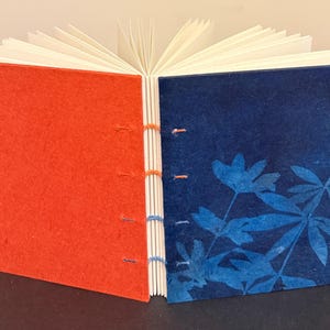 May include: Open handmade journal with a red cover on the left and a blue cover on the right. The blue cover features a floral design. The pages are white and the binding is hand-stitched with orange and blue thread.