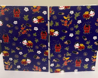 May include: Two small blue notebooks with a repeating pattern of red and gold Japanese characters, red and gold mythical creatures, and gold flowers. The notebooks are bound together with a black string.