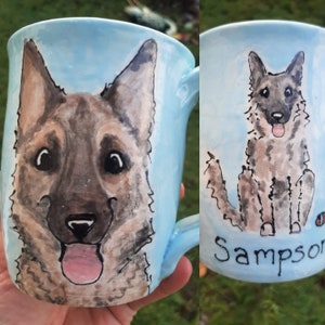 May include: A light blue ceramic mug with a hand-painted portrait of a dog. The dog has brown and black fur, a pink tongue sticking out, and black eyes. The mug has the name "Sampson" written on the bottom.