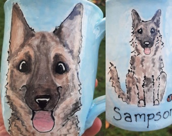 Puede incluir: Una taza de cerámica azul claro con un retrato pintado a mano de un perro. El perro tiene pelo marrón y negro, una lengua rosa que sobresale y ojos negros. La taza tiene el nombre "Sampson" escrito en la parte inferior.