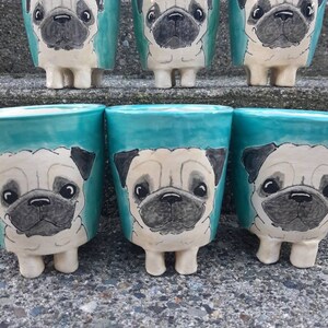 Puede incluir: Seis tazas de cer&aacute;mica de color turquesa con caras de perro pug pintadas. Las tazas tienen peque&ntilde;os pies y est&aacute;n dispuestas en una fila sobre una superficie de piedra gris.