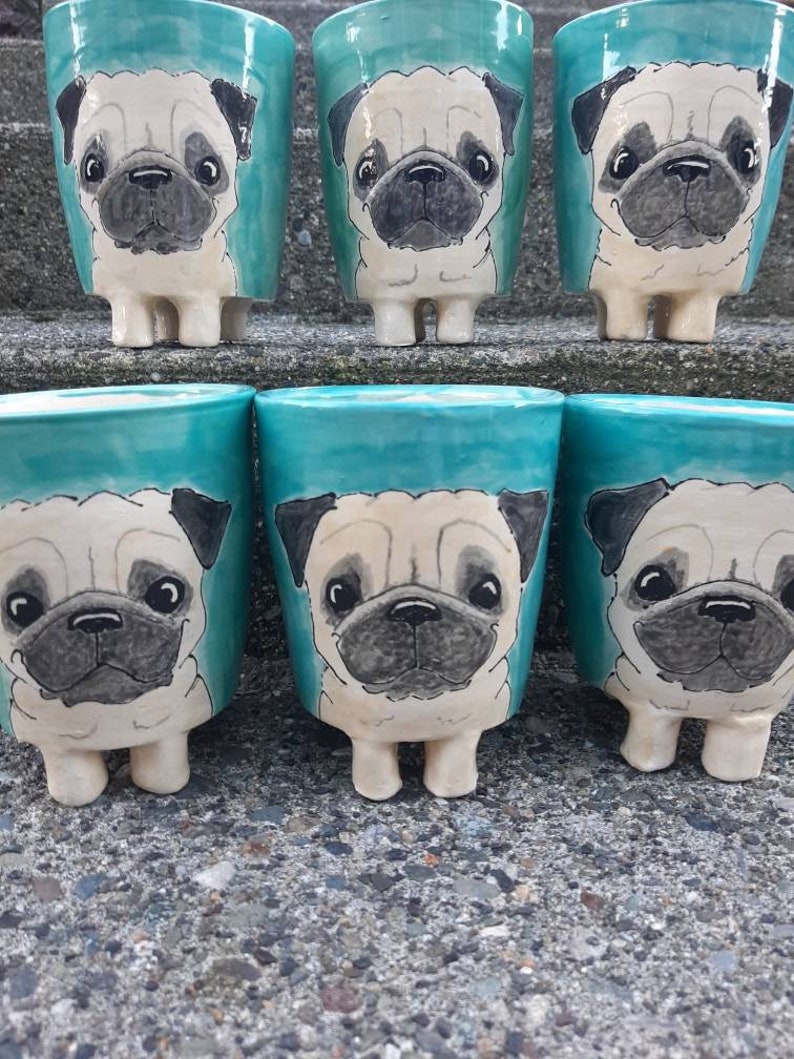Puede incluir: Seis tazas de cer&aacute;mica de color turquesa con caras de perro pug pintadas. Las tazas tienen peque&ntilde;os pies y est&aacute;n dispuestas en una fila sobre una superficie de piedra gris.