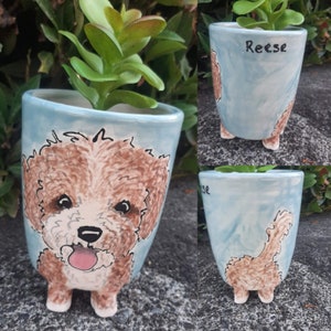 May include: A light blue ceramic planter with a painted image of a brown and white dog with a pink tongue sticking out. The planter has four small feet and the name "Reese" is written on the side.