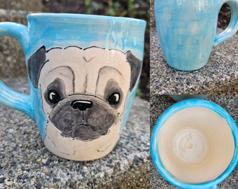 Puede incluir: Una taza de cerámica azul claro con una cara de carlino pintada a mano. La taza tiene un esmalte blanco y un esmalte azul claro.