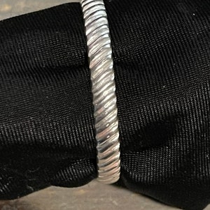 Peut inclure: Une bague argentée avec un motif torsadé en forme de corde. La bague est faite d'un matériau métallique brillant et est posée sur un fond sombre. La surface de la bague présente une série de rainures diagonales, créant un effet de spirale.
