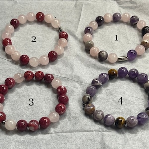 May include: Four beaded bracelets with different gemstone combinations. Bracelet 1 features purple, pink, and gray beads. Bracelet 2 has pink and red beads. Bracelet 3 is red and pink. Bracelet 4 has purple, brown, and yellow beads.