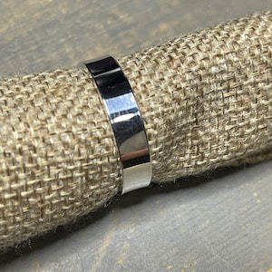 May include: A polished silver ring with a flat, rectangular band. The ring is shown on a textured, tan surface. The ring's reflective surface catches the light, highlighting its smooth, clean lines. A simple, elegant piece of jewelry.