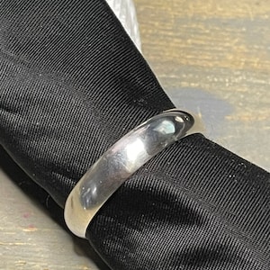 Puede incluir: Un anillo de plata pulida con un diseño liso y redondeado. El anillo se encuentra sobre un fondo de tela negra, creando un contraste que resalta su superficie reflectante. La elegancia sencilla del anillo lo convierte en un accesorio versátil.