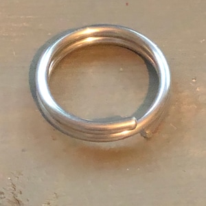 Peut inclure: Un porte-clés argenté avec un design à triple boucle. Le porte-clés est en métal et de forme circulaire. Le porte-clés est posé sur une surface brun clair.