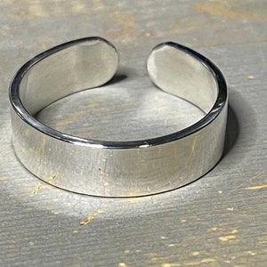 May include: A silver adjustable ring with a smooth, flat surface. The ring is open on one side and can be adjusted to fit different finger sizes.