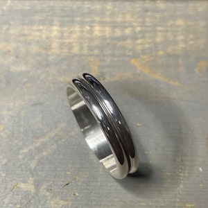 May include: A polished silver ring with a double-grooved design. The ring has a smooth, reflective surface, and the grooves run parallel around the band. The ring is shown on a gray surface.