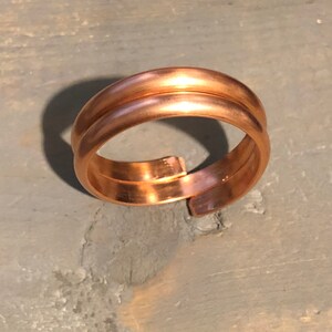 Peut inclure: Une bague de couleur cuivre avec un design à double bande. La bague a une finition lisse et polie et un design ouvert et réglable. La bague est présentée sur une surface grise texturée.