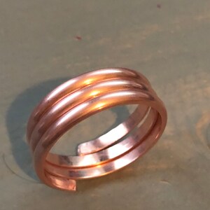 Peut inclure: Une bague en cuivre à trois anneaux. Les anneaux sont lisses et polis. La bague est présentée sur un fond vert clair.