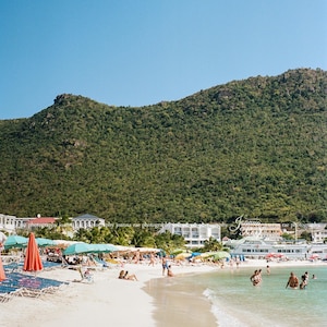 May include: A beach scene with white sand, clear blue water, and a lush green hillside in the background. There are people swimming and relaxing on the beach, and several colorful umbrellas are set up.