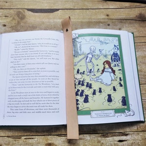 Puede incluir: Una ilustración vintage del libro "El Maravilloso Mago de Oz" de L. Frank Baum. La ilustración representa a Dorothy, el Hombre de Hojalata y el Espantapájaros de pie en un campo de ratones. Los ratones son todos negros y blancos y están dispuestos en un círculo alrededor de los tres personajes. El Hombre de Hojalata sostiene un pájaro en su mano y el Espantapájaros sostiene una escoba. Dorothy está sentada en el suelo, mirando al Hombre de Hojalata.
