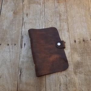 May include: A brown leather journal with a snap closure. The journal is lying on a wooden surface.