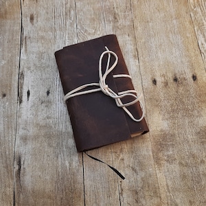 May include: A brown leather-bound journal with a white string closure. The journal is closed and tied with the string. The journal is sitting on a wooden surface.