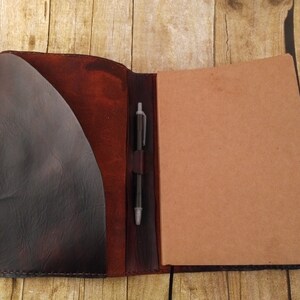 May include: A brown leather journal with a pen holder and a flap pocket. The journal is open to reveal blank pages.