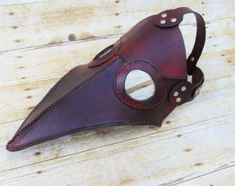 Könnte beinhalten: Eine braune Ledermaske eines Pestarztes mit einem langen Schnabel und zwei runden Augenlöchern. Die Maske hat ein Band, das um den Kopf geht.