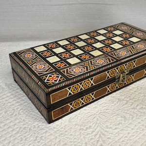 May include: A rectangular wooden box with a chessboard design on the lid. The box is brown with intricate inlaid patterns in various colors, including orange, white, and black. A gold-colored clasp secures the box.