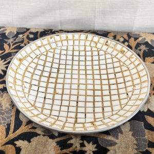May include: A round, white plate with a gold grid pattern and a silver rim. The plate has a mosaic-like design and is set against a floral patterned background. This decorative plate is suitable for serving food or as a decorative item.