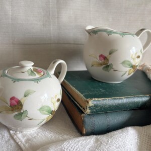 May include: A white porcelain teapot with a matching creamer. Both pieces are decorated with pink and yellow floral designs and green leaves. The teapot has a lid and a handle. The creamer sits on a stack of vintage green books.