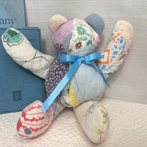 May include: A stuffed teddy bear constructed from various patchwork fabric scraps in multiple colors and patterns, including floral, gingham, and geometric prints. The bear features a bright blue satin ribbon tied around its neck.