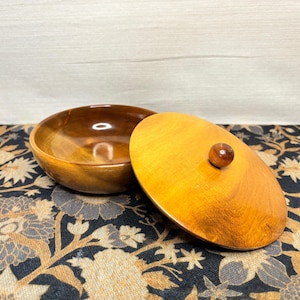 May include: A wooden bowl with a matching lid. The bowl is open, revealing a glossy interior. The lid is off to the side, showing a small wooden knob. The wood has a warm, golden-brown hue.