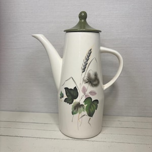May include: A white ceramic coffee pot with a green lid and handle. The pot is decorated with a floral and wheat design in shades of green, gray, and pink. The spout is long and slender.