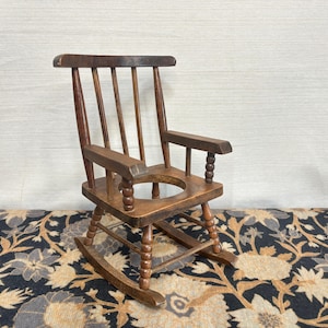 May include: A small, antique wooden rocking chair with a dark brown finish. The chair has a round seat, armrests, and a slatted back. The chair is on a patterned rug.