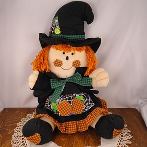 May include: A plush doll featuring orange yarn hair, a black witch hat with a spiderweb print, and a black dress with a green gingham bow. The doll has round orange gingham cheeks, a black bodice, and a skirt with a spiderweb and orange gingham trim.