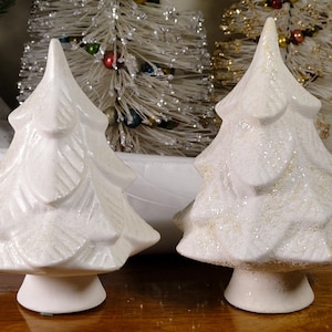 May include: Two white ceramic Christmas tree figurines. One tree has a textured surface, while the other is covered in glitter. Both have a classic tree shape with a conical top and a round base. The trees are decorative holiday accents.
