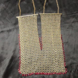 Op de afbeelding: Een gouden ketting met een delicaat ontwerp met kleine witte kralen en rode druppelvormige stenen. De ketting bestaat uit twee afzonderlijke strengen die aan de bovenkant met elkaar verbonden zijn.