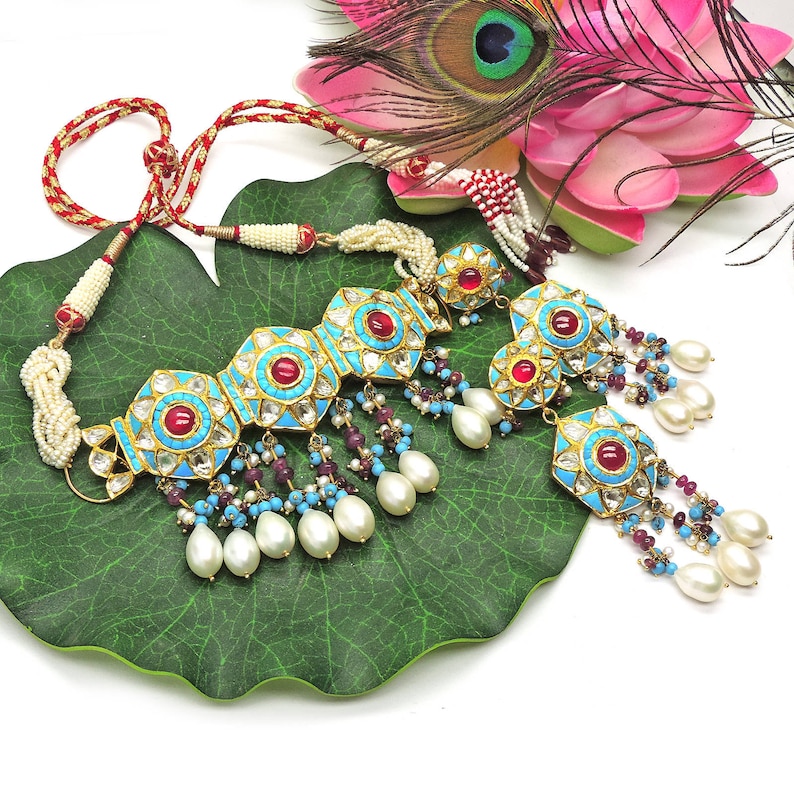 May include: A turquoise and gold necklace with red and white stones, featuring a central pendant with a floral design and dangling pearl and turquoise beads.