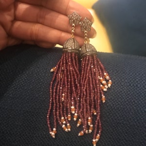 Puede incluir: Un par de pendientes colgantes con una parte superior de diamante en forma de estrella y una larga borla de cuentas color burdeos. La borla está adornada con pequeñas cuentas naranjas y blancas.