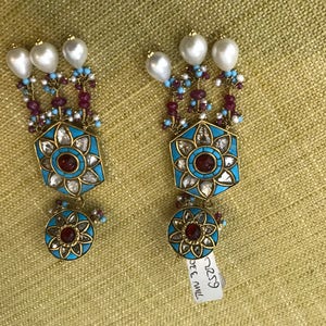 May include: A pair of long, dangling earrings with a blue and gold design. The earrings feature a hexagonal pendant with a red stone in the center, surrounded by smaller white stones. The pendant is attached to a chain of blue and red beads, and the chain is topped with three white pearls.