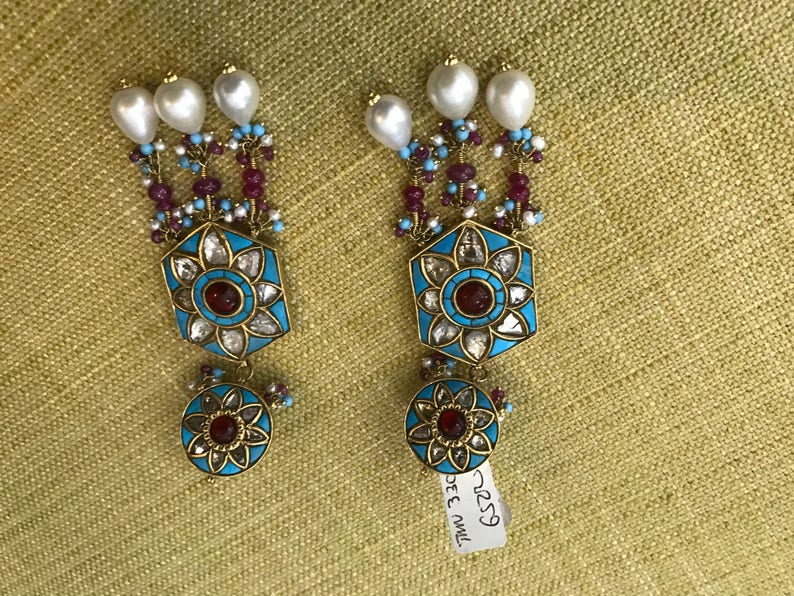 May include: A pair of long, dangling earrings with a blue and gold design. The earrings feature a hexagonal pendant with a red stone in the center, surrounded by smaller white stones. The pendant is attached to a chain of blue and red beads, and the chain is topped with three white pearls.