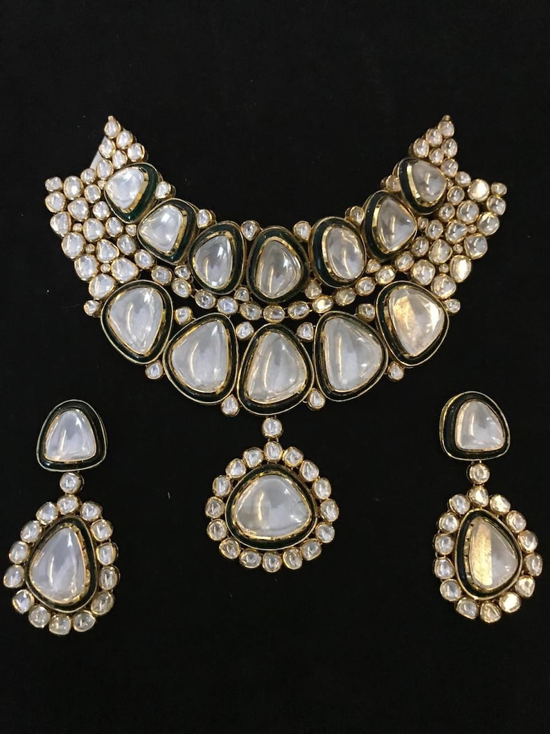Pu&ograve; includere: Un set di collana e orecchini in oro con grandi pietre trasparenti e sfaccettate incastonate in un bordo di smalto verde. Le pietre sono circondate da pietre pi&ugrave; piccole e trasparenti.