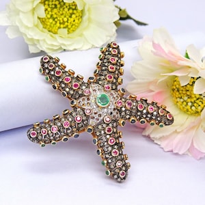 Puede incluir: Un broche de estrella de mar de oro con una gran piedra preciosa verde esmeralda en el centro, rodeada de piedras preciosas más pequeñas de color rosa, rojo y blanco. El broche se coloca sobre un fondo blanco.