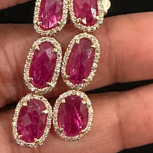 Op de afbeelding: Een paar oorhangers met drie ovale roze edelstenen, gezet in goud met een halo van kleinere diamanten.
