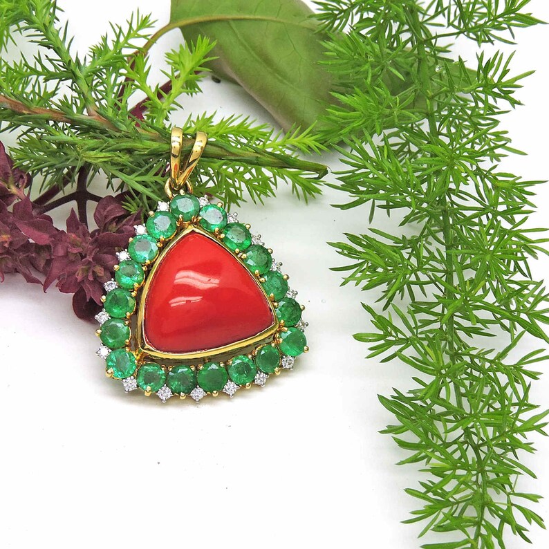 May include: A triangular pendant featuring a large red coral gemstone, encircled by a halo of green emeralds and small diamonds.