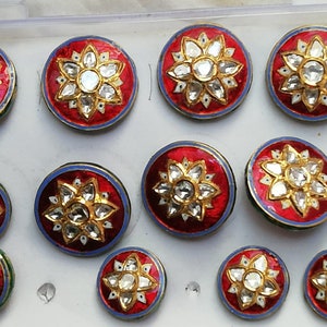 May include: Ten round, red and blue buttons with a white and gold flower design in the centre. Each button has a white centre with a clear stone in the middle. The buttons are arranged in a grid pattern.