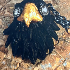 May include: A black leather eagle pendant with a gold beak and rhinestone eyes. The pendant is attached to a black chain.