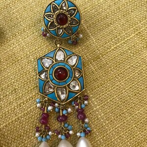 May include: A pair of gold earrings with a turquoise and white enamel design. The earrings feature a red gemstone in the center of each flower design and dangle with pearls and red and blue beads.
