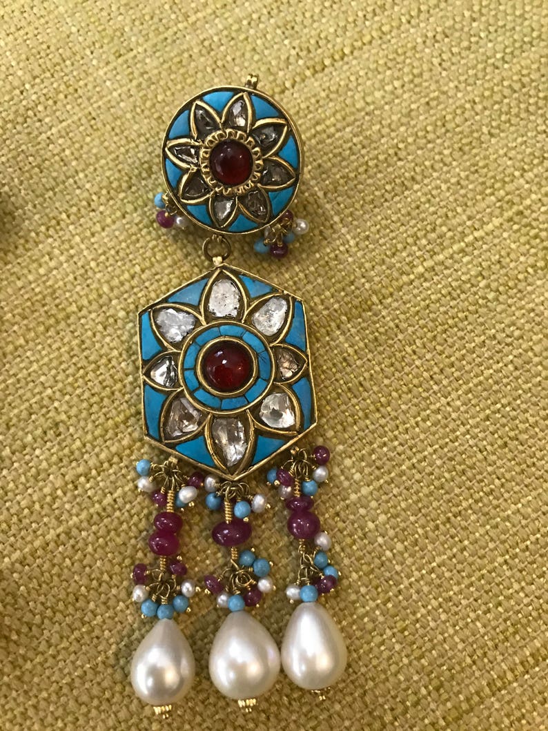 May include: A pair of gold earrings with a turquoise and white enamel design. The earrings feature a red gemstone in the center of each flower design and dangle with pearls and red and blue beads.