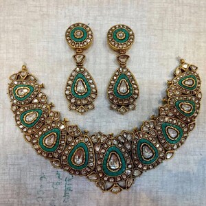 May include: A gold necklace and earrings set with a teardrop design. The necklace is made up of multiple teardrop-shaped pendants with green and white stones. The earrings are also teardrop-shaped with green and white stones.