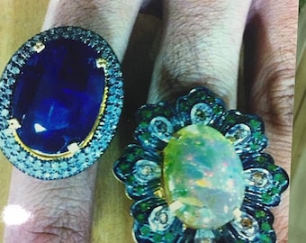 May include: Two rings with large gemstones. The ring on the left has a blue sapphire surrounded by a halo of smaller diamonds. The ring on the right has a large opal surrounded by a halo of smaller diamonds and green stones.