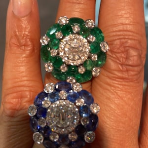 May include: Two rings with a cluster of gemstones set in a circular pattern. The top ring features a large emerald green stone surrounded by smaller emeralds and diamonds. The bottom ring features a large sapphire blue stone surrounded by smaller sapphires and diamonds.