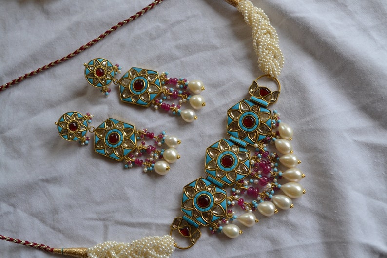 May include: A gold necklace and earrings set with turquoise and red stones, white pearls, and pink beads. The necklace has a choker style with a decorative pendant.