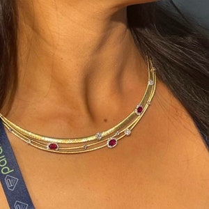 Puede incluir: Un collar de oro de tres capas. El collar presenta pequeñas piedras preciosas rojas y diamantes transparentes.